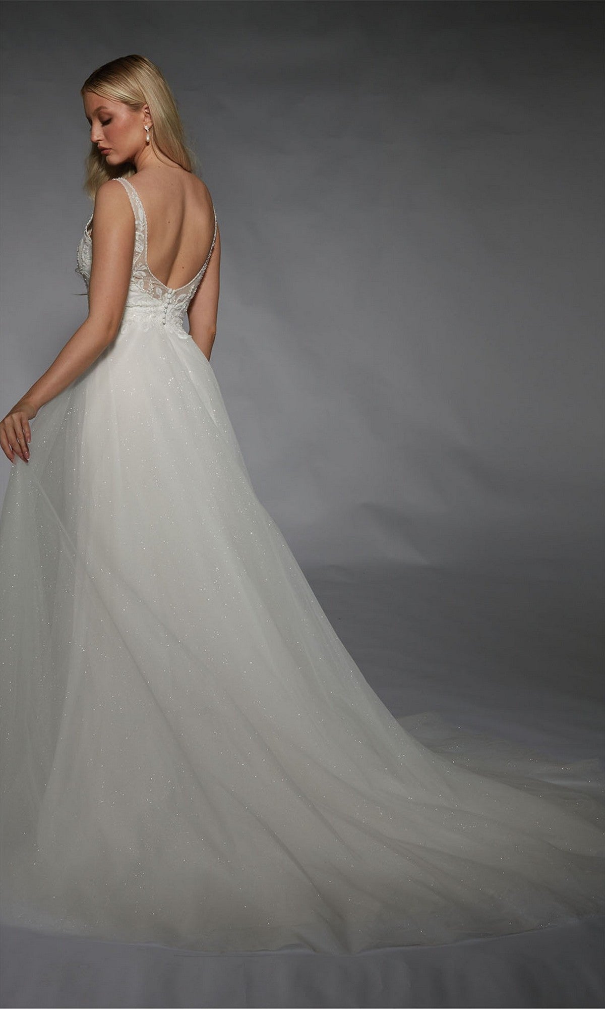 A woman poses against a plain gray background, wearing the Long White Dress: Alyce 7125—a sleeveless white gown with an open back and a long, flowing train.